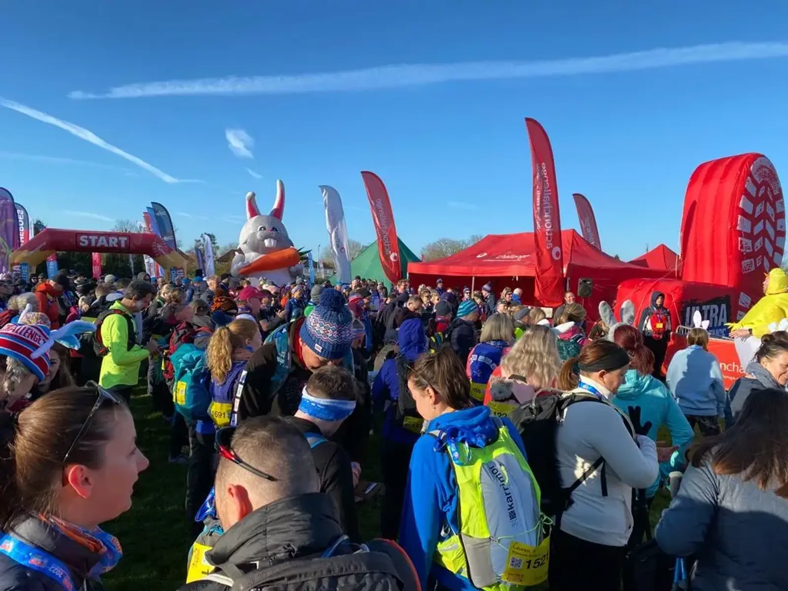 A large crowd of runners gathers at the start line of a race event. People wear colorful athletic gear and backpacks. Red tents and flags are visible in the background under a clear blue sky.