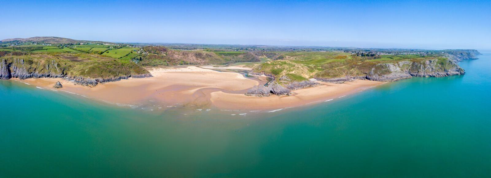 Aerial view of a wide, sandy beach surrounded by green cliffs and turquoise water. The beach is nestled between two rocky headlands, with lush countryside visible in the background under a clear blue sky.