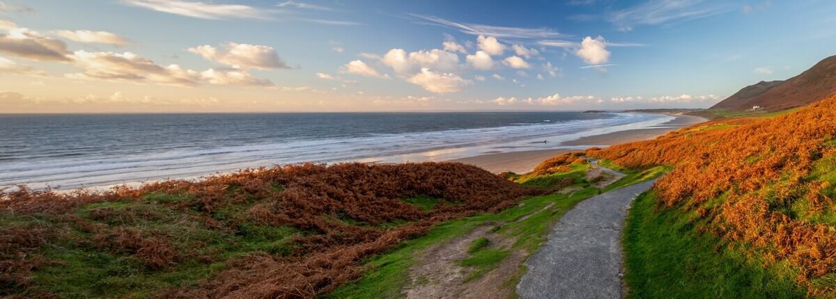 A scenic view of a coastal path leading to a sandy beach, with the ocean on the left and hills on the right. The foreground features green and brown vegetation under a blue sky with scattered clouds at sunset.