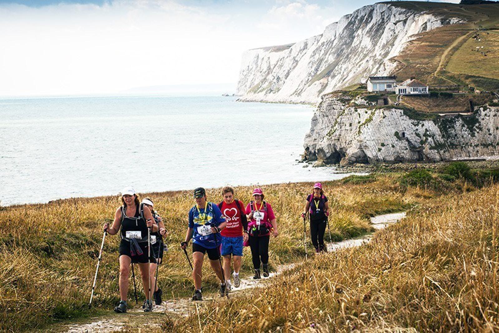 A group of people hiking along a coastal path with tall cliffs and the ocean in the background. The trail is surrounded by grassy fields, and the hikers are equipped with backpacks and trekking poles. The day is clear and bright.