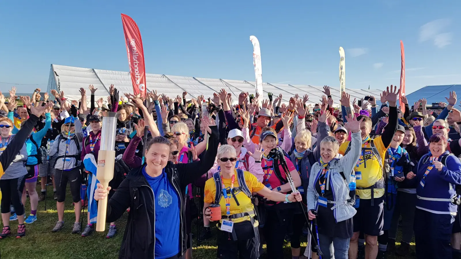 A large group of people in colorful outdoor attire are gathered together, smiling and raising their hands in celebration. They appear to be at an outdoor event near tents, with several flags in the background against a clear blue sky.