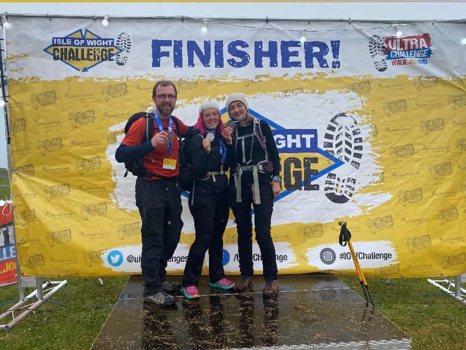 Three people in hiking gear pose in front of a "FINISHER!" backdrop for the Isle of Wight Challenge. They stand on a wet surface, holding medals, and giving thumbs up. A hiking pole rests on the ground beside them.