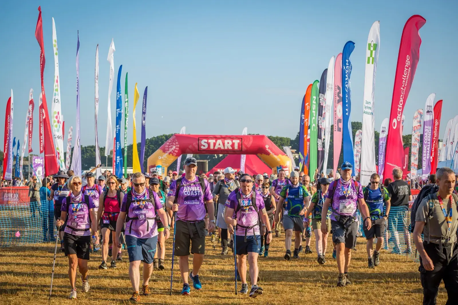 A group of people wearing colorful event shirts and carrying backpacks participate in an outdoor charity walk. They are walking past a start line, surrounded by flags and a clear blue sky overhead.