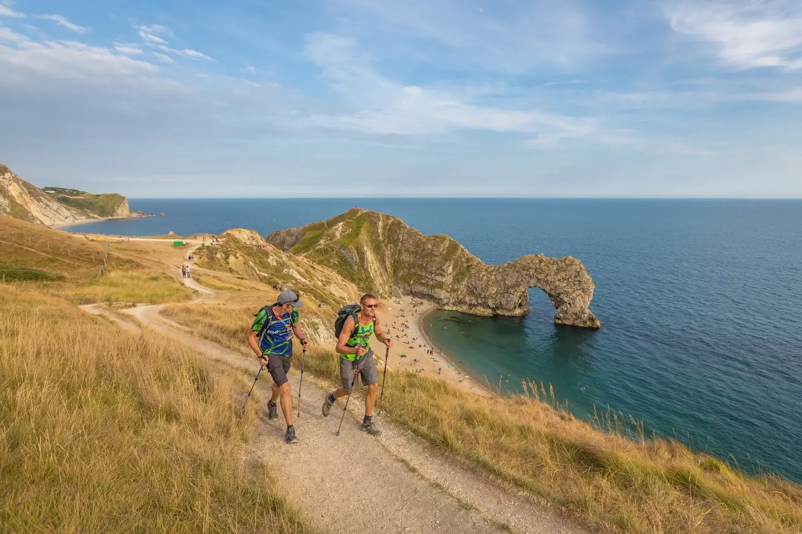 Two hikers with backpacks and walking poles trek along a coastal path overlooking Durdle Door, a natural limestone arch in Dorset, England. The blue sea, green grass, and clear sky create a picturesque landscape.