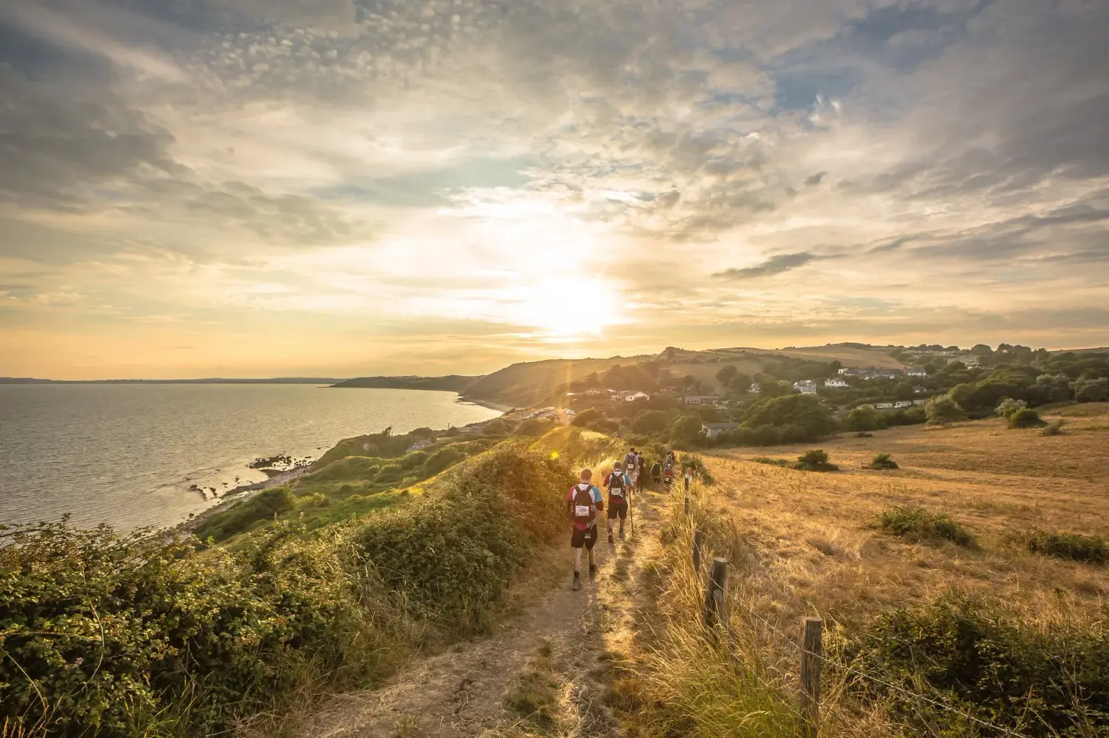A group of people hikes along a coastal trail at sunset. The path is surrounded by grassy fields and shrubs, with the ocean on the left and hills in the distance under a partly cloudy sky. The sun is setting on the horizon.