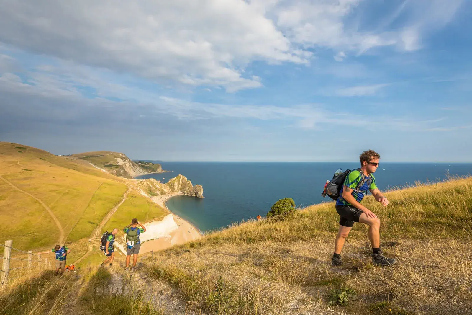 Hikers traverse a grassy hill overlooking the coastline, with a blue sky above and a curving sandy beach below. A dramatic cliff extends into the sea, creating a striking natural vista.