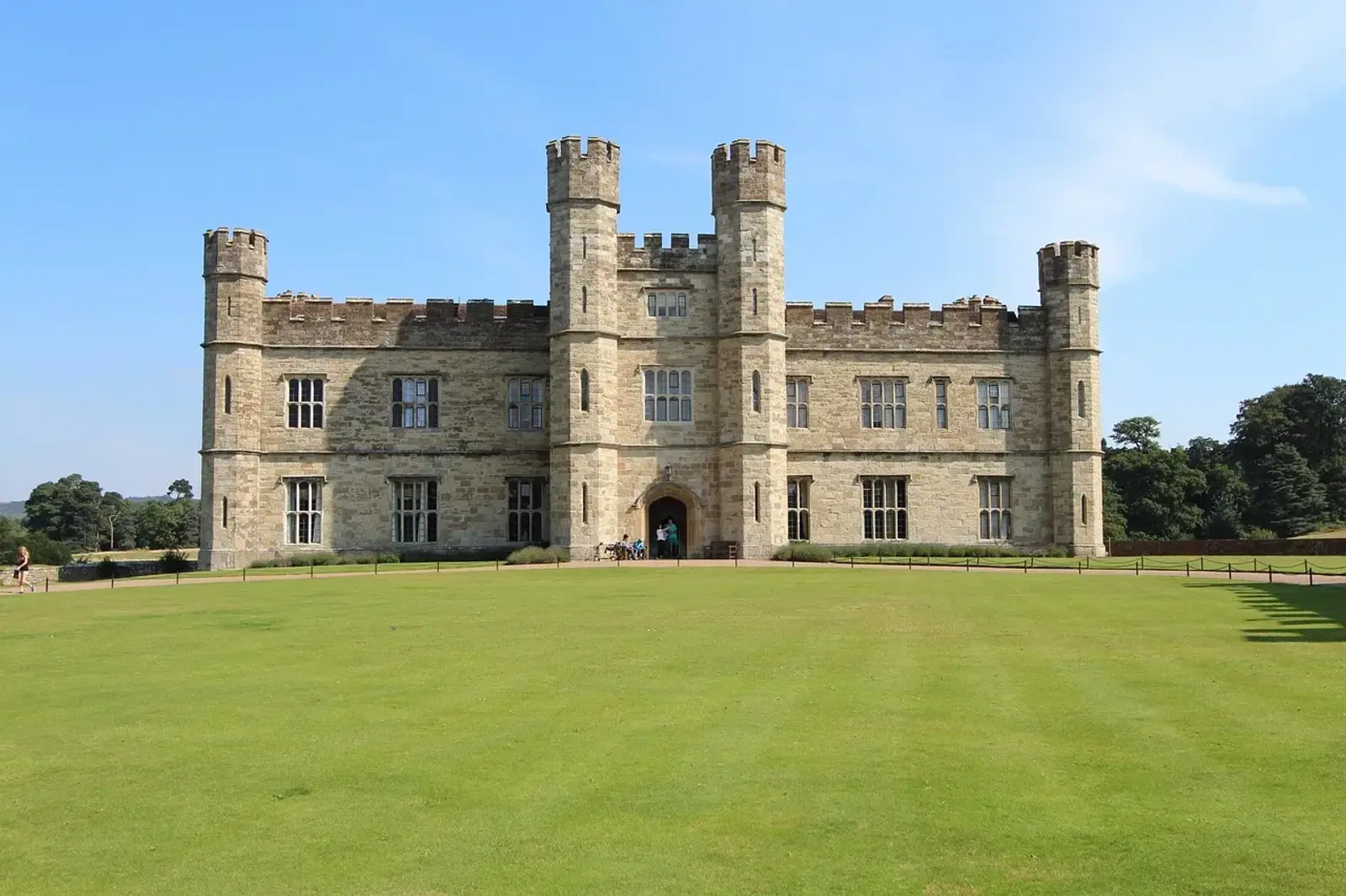 A large, historic stone castle with four tall towers sits against a clear blue sky. The building is surrounded by a well-kept green lawn, with pathways leading up to its entrance. Trees are visible in the background.