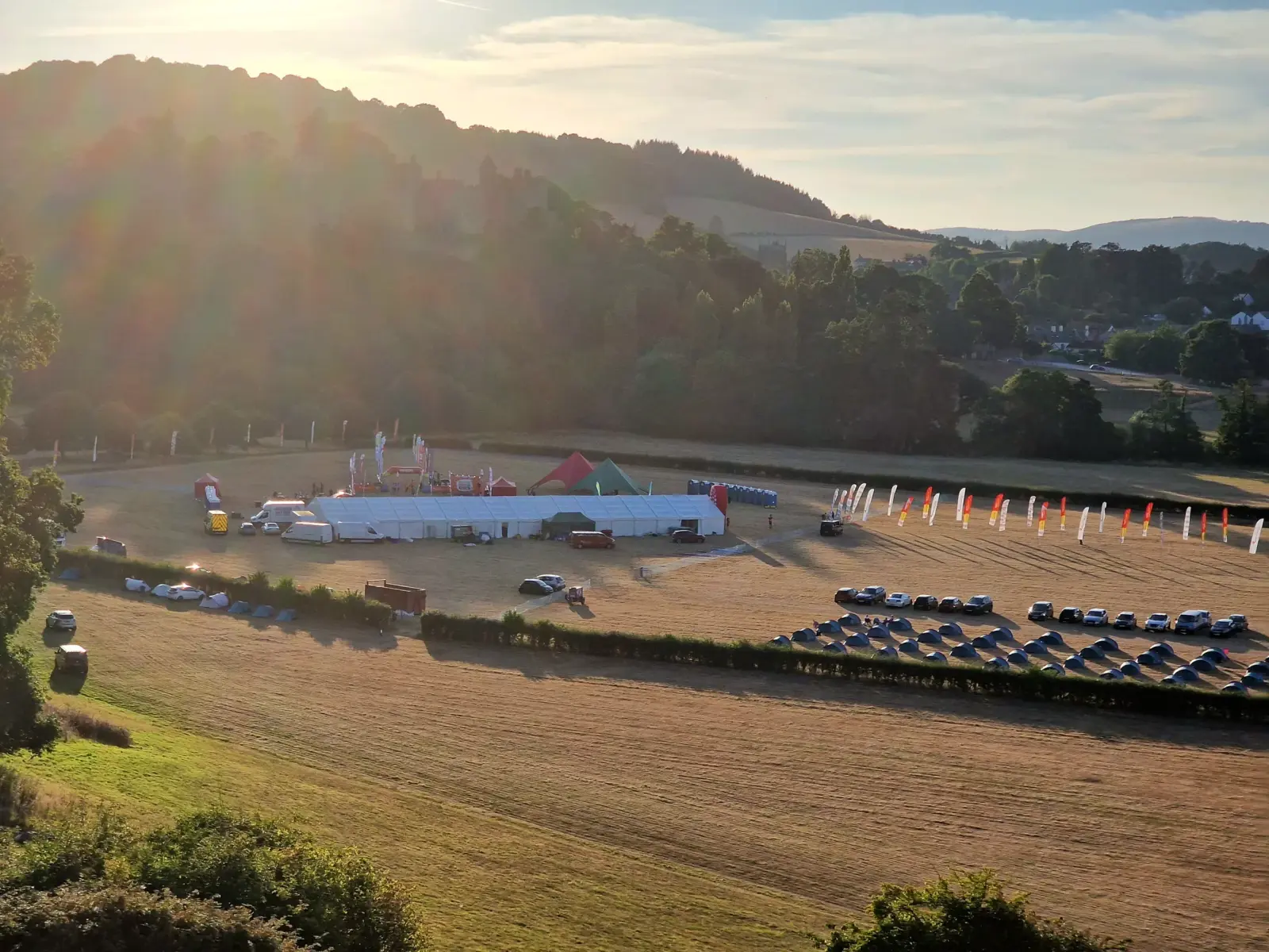 A scenic view of a rural fairground with tents and colorful flags on a sunny day. Several vehicles are parked in a field, surrounded by lush greenery and hills in the background. The sunlight casts a warm glow over the landscape.