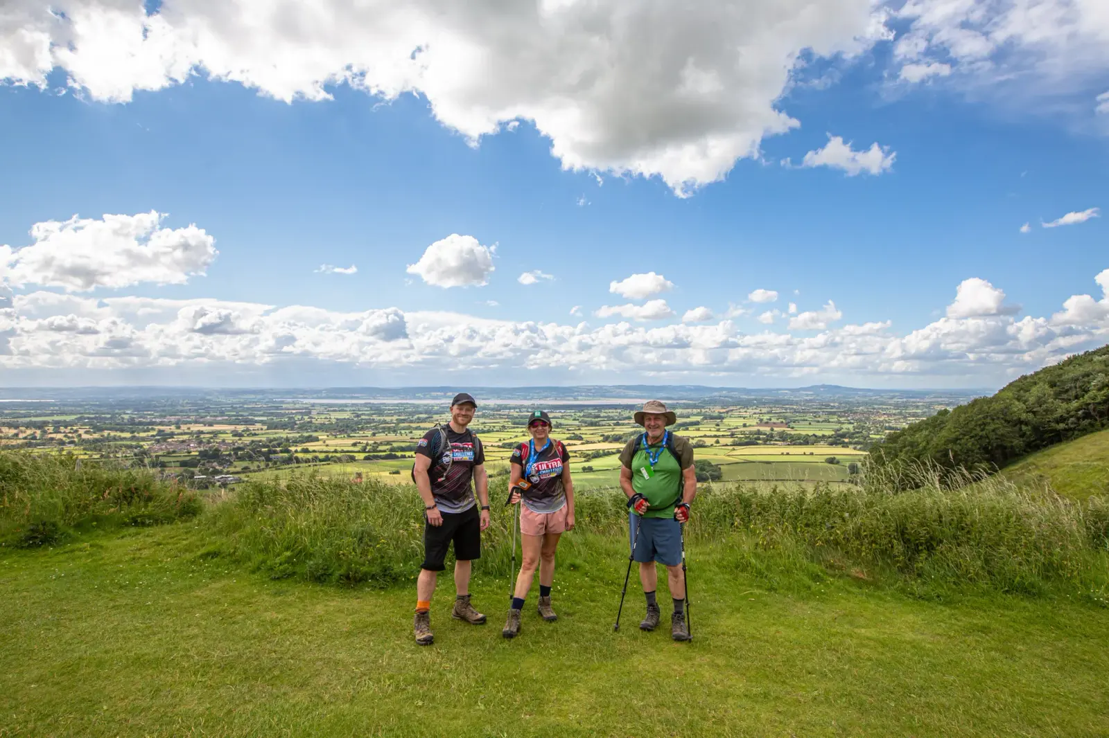 Three people stand on a grassy hilltop with a scenic view of a countryside landscape under a partly cloudy sky. They are dressed in casual outdoor attire, with one person holding walking sticks. Verdant fields stretch out in the background.