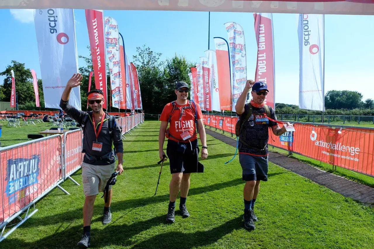 Three men wave as they walk under a finish line banner at an outdoor event. They carry trekking poles and wear sporty outfits. Red and white flags and barriers with event branding line the grassy path. Sunny weather highlights the vibrant scene.