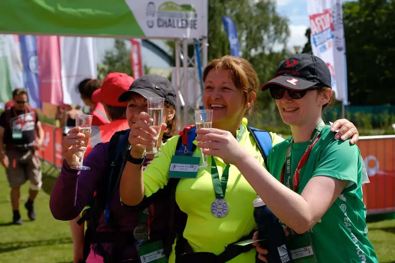 Three women wearing sports attire and medals cheer with plastic cups in a sunny outdoor event setting. Banners and other participants are visible in the background.