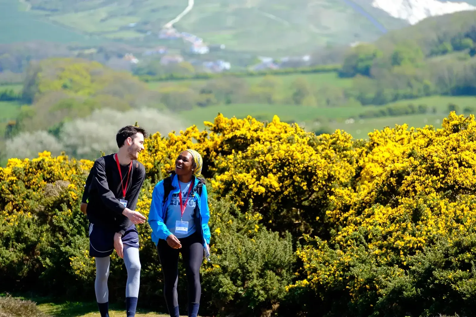 Two people hike along a trail bordered by vibrant yellow flowers. They are smiling at each other, wearing sporty clothing and event badges. The background features lush green hills and a clear blue sky.