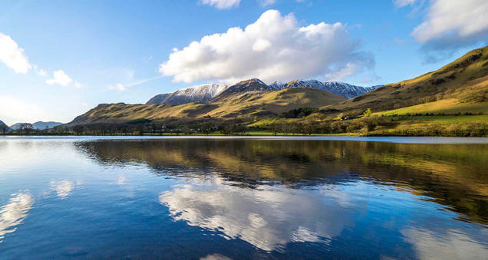 A serene lake reflecting fluffy white clouds and distant snow-capped mountains under a blue sky. The surrounding rolling hills are covered in green grass, creating a tranquil and picturesque landscape.