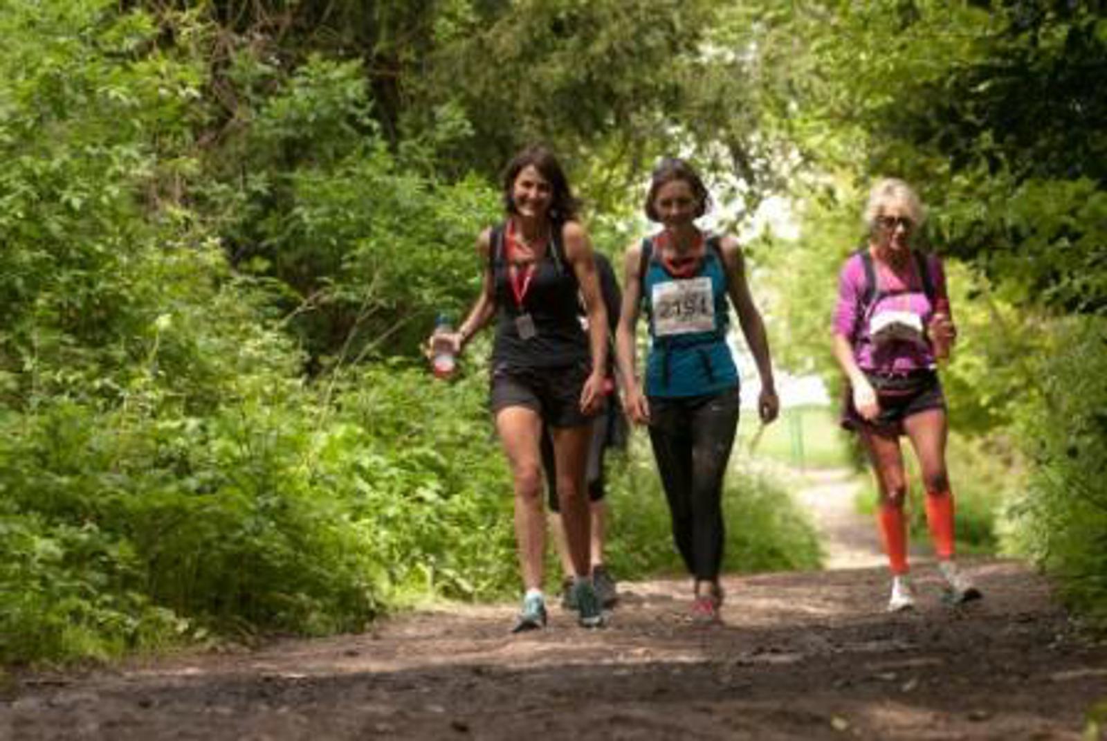 Three women walking on a forest trail, wearing race numbers and medals. They are dressed in sportswear, and the path is surrounded by lush green foliage.