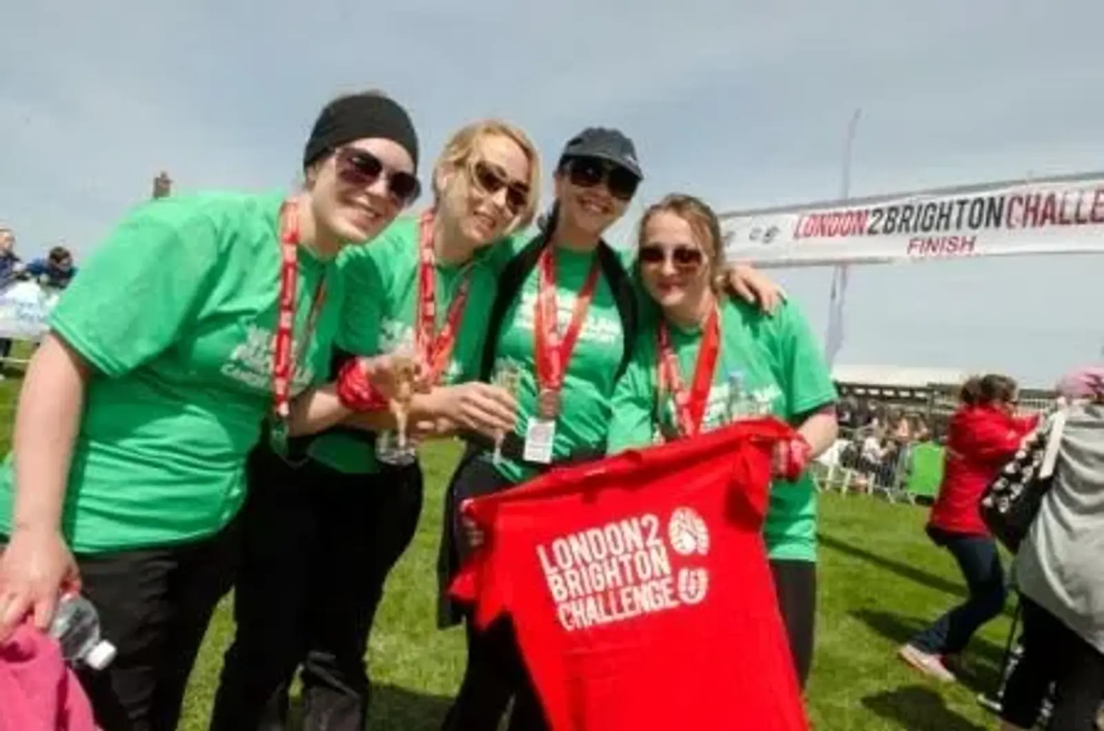 Four people wearing green "London 2 Brighton Challenge" shirts celebrate at the finish line, holding medals and a red event shirt. They are smiling under a sunny sky with a banner in the background.