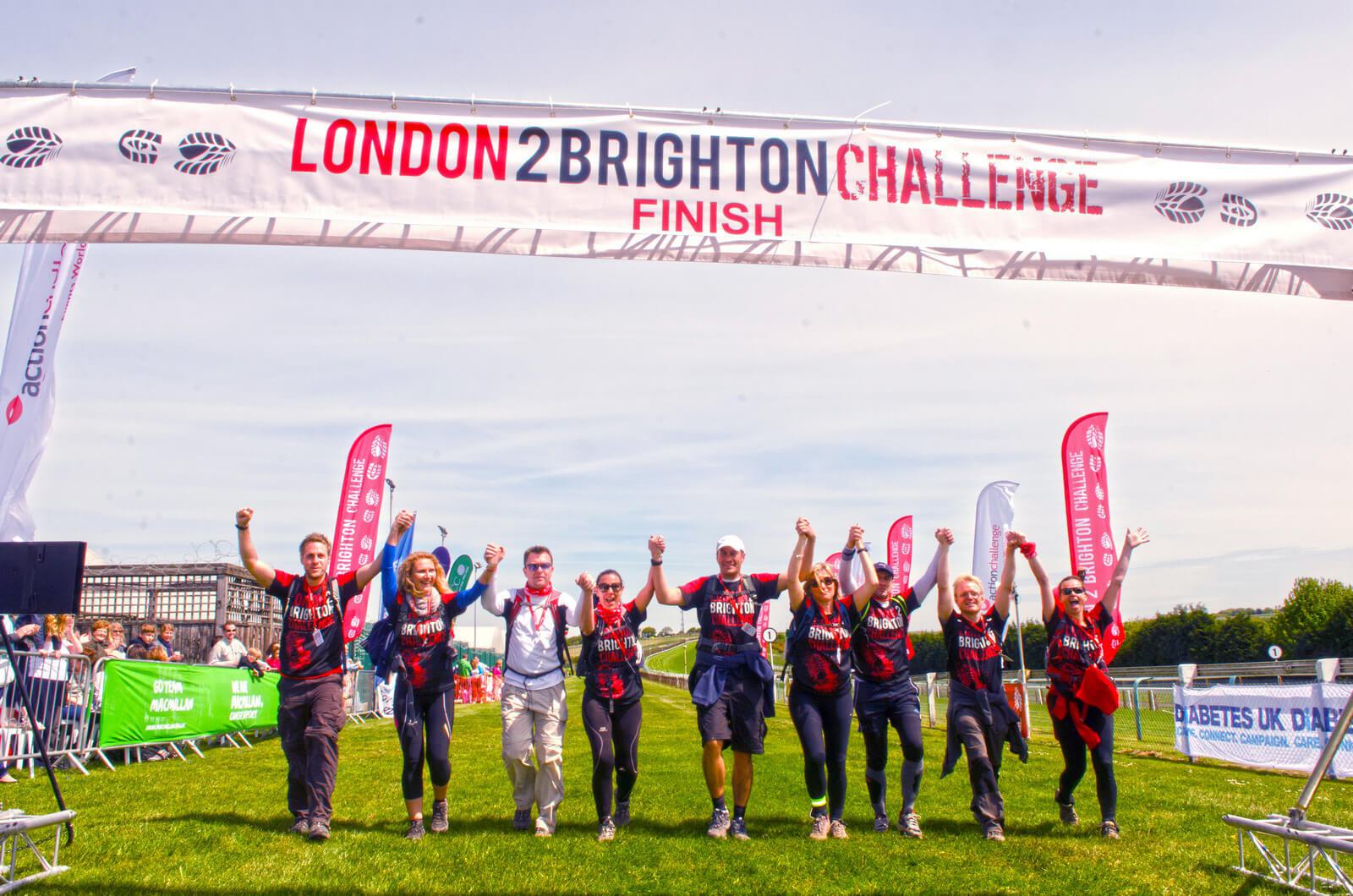 A group of seven people celebrating as they cross the finish line of the London 2 Brighton Challenge under a large banner. They're wearing matching outfits and raising their arms triumphantly. Flags and spectators are visible in the background.