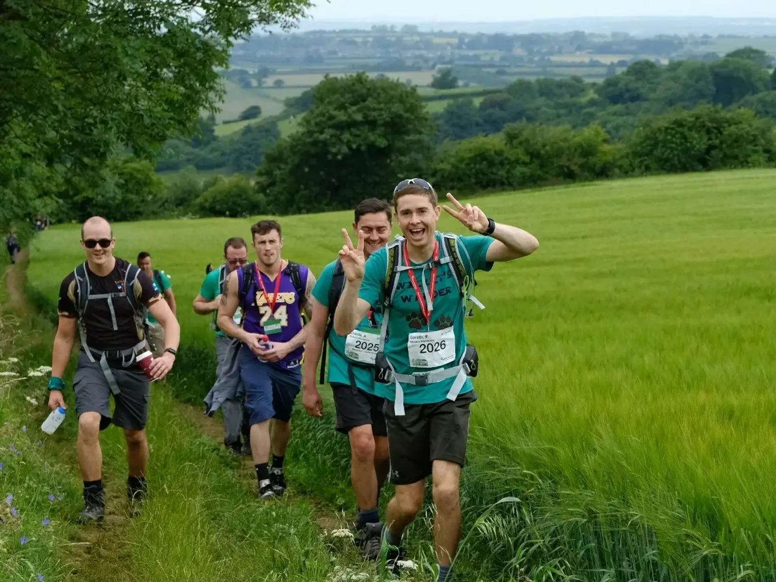 A group of five hikers walking through a lush green field on a countryside trail. One person in front is smiling and gesturing peace signs. The landscape includes rolling hills and distant trees under a cloudy sky.