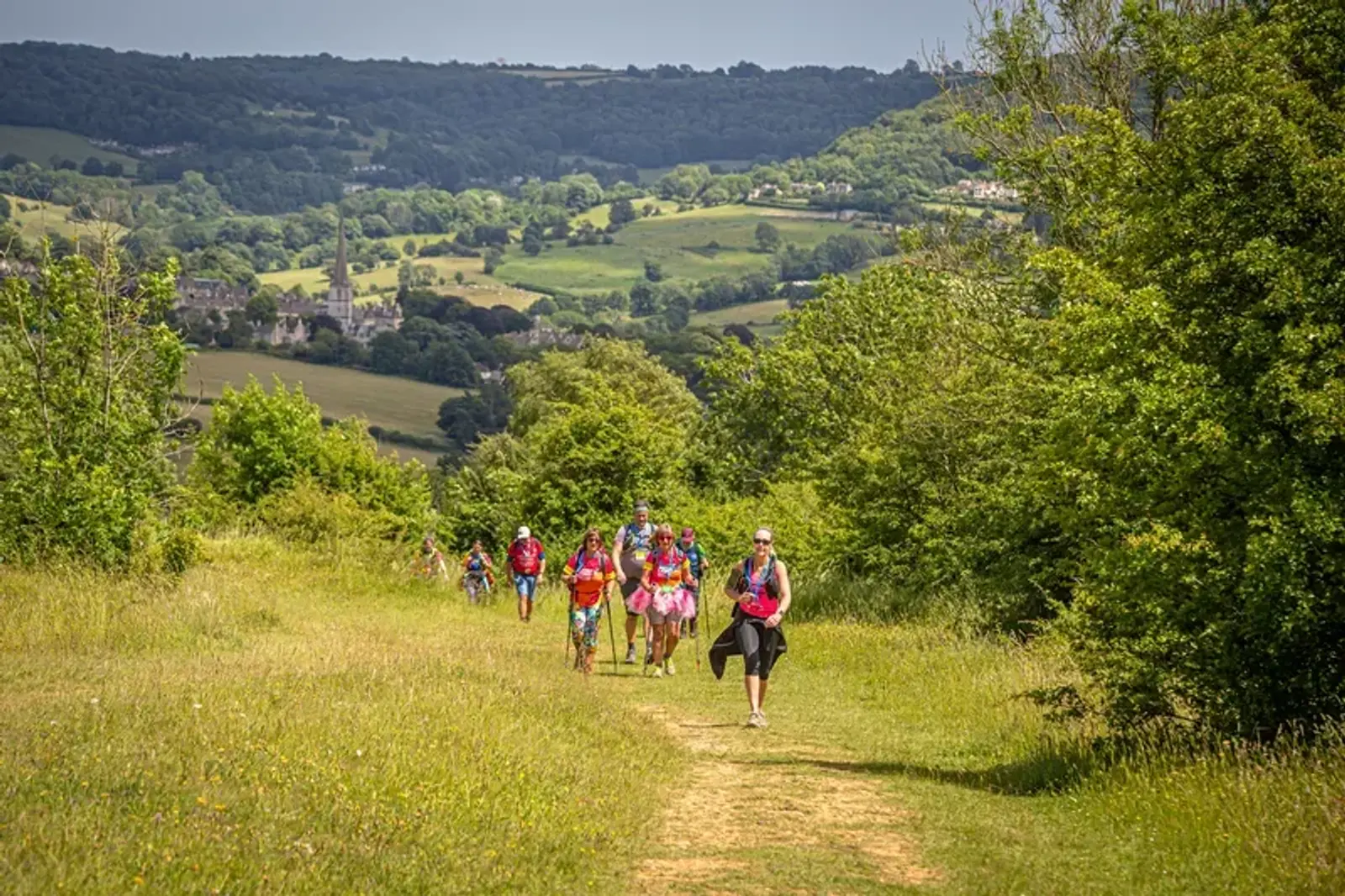 A group of people hiking up a grassy trail in a lush, green countryside. Trees line the path, and a scenic village is visible in the valley below. It appears to be a sunny day, highlighting the vibrant landscape.