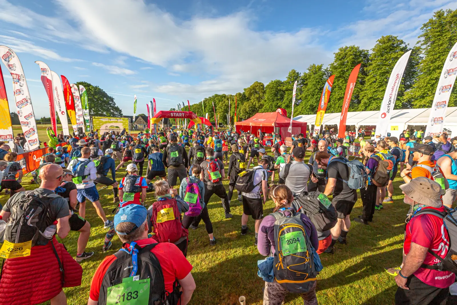 A large group of hikers gathers at a starting line for a race in a grassy area. They wear colorful shirts and backpacks with numbers. Flags and tents are set up in the background under a partly cloudy sky.