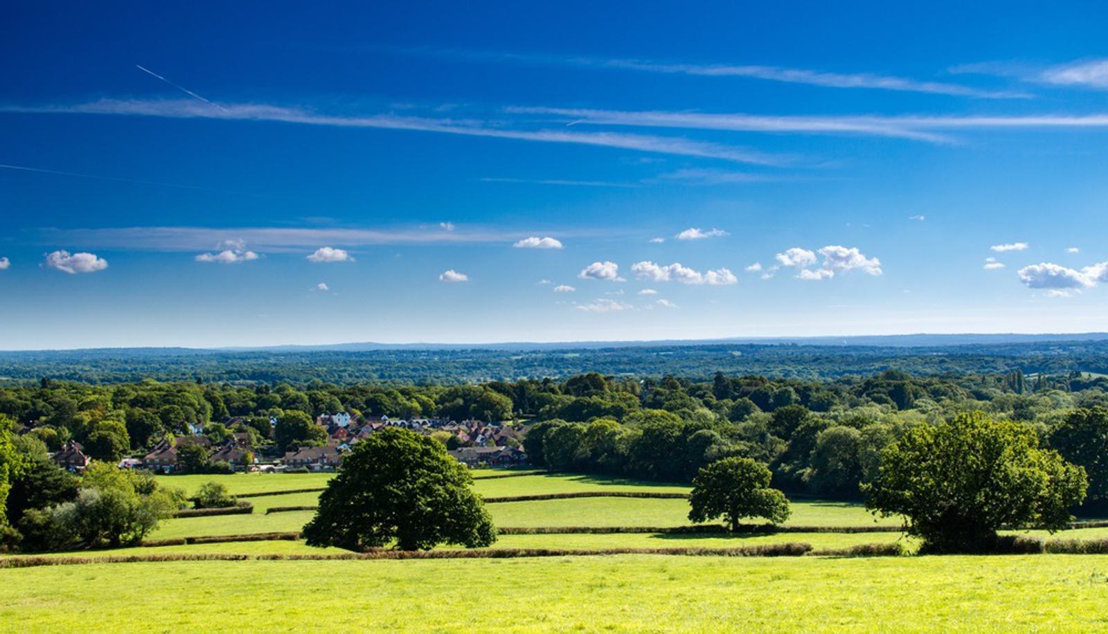 Expansive rural landscape featuring green fields, scattered trees, and a small village nestled among lush greenery. The horizon stretches under a bright blue sky with thin clouds.