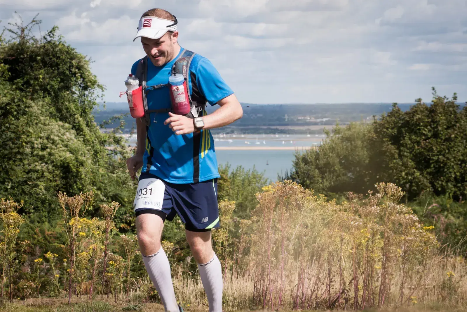 A man is running outdoors on a trail, wearing a blue shirt, black shorts, white compression socks, and a hydration pack. He has a race bib pinned to his shorts. The background shows greenery and a distant body of water under a partly cloudy sky.