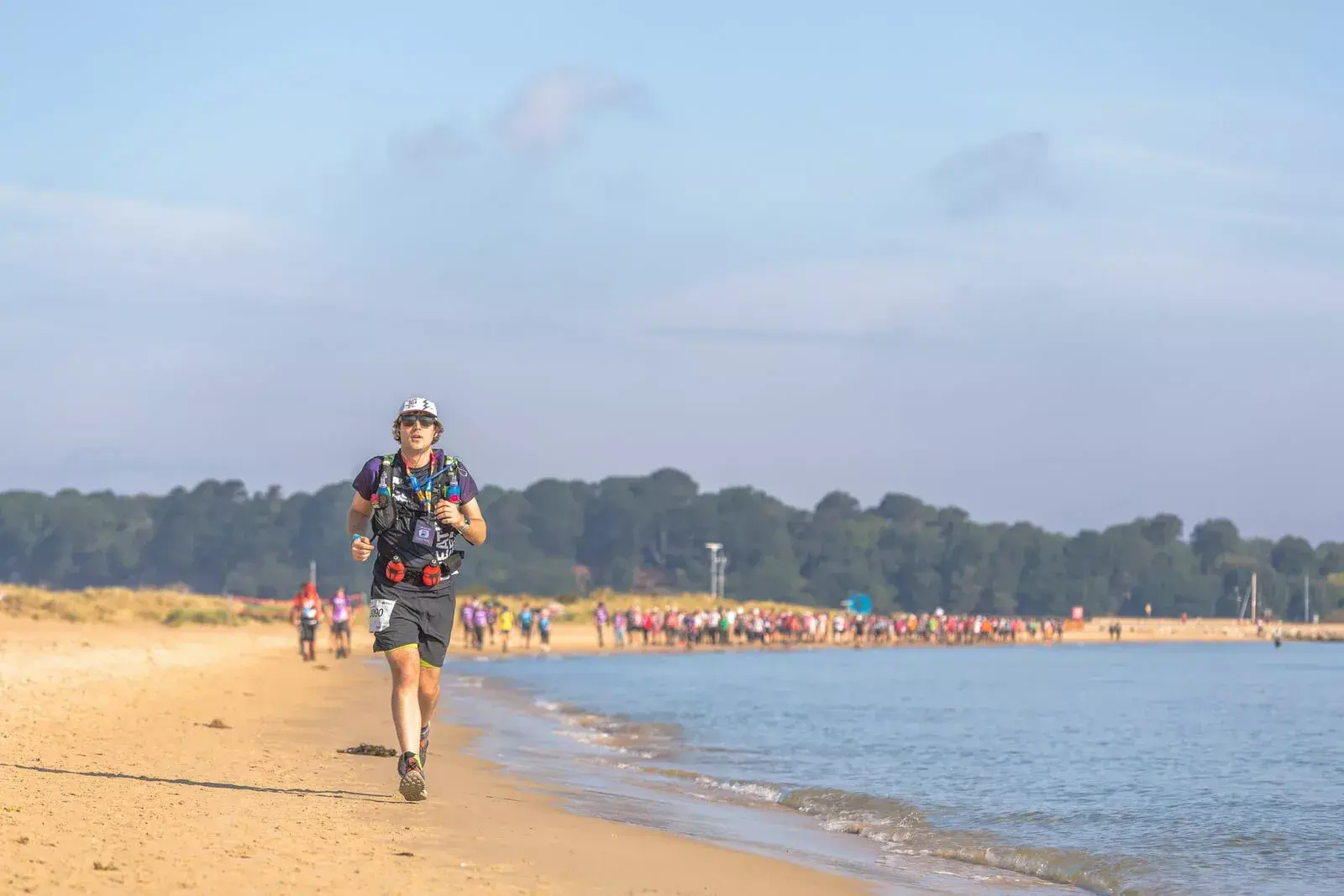 A person in athletic gear runs along a sandy beach with a calm sea to the right. A crowd of people can be seen in the distance under a clear blue sky, with a forested area in the background.