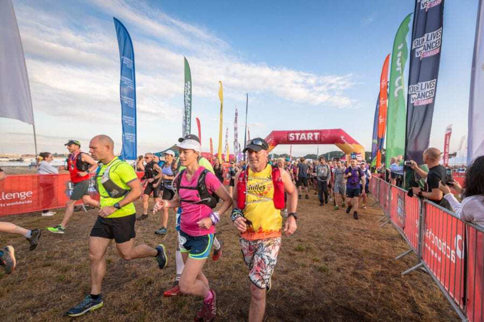 A group of runners in colorful athletic gear participate in a race, passing under a red start arch. Bright flags and banners line the path, with onlookers cheering them on. The sky is clear, suggesting it's a sunny day.
