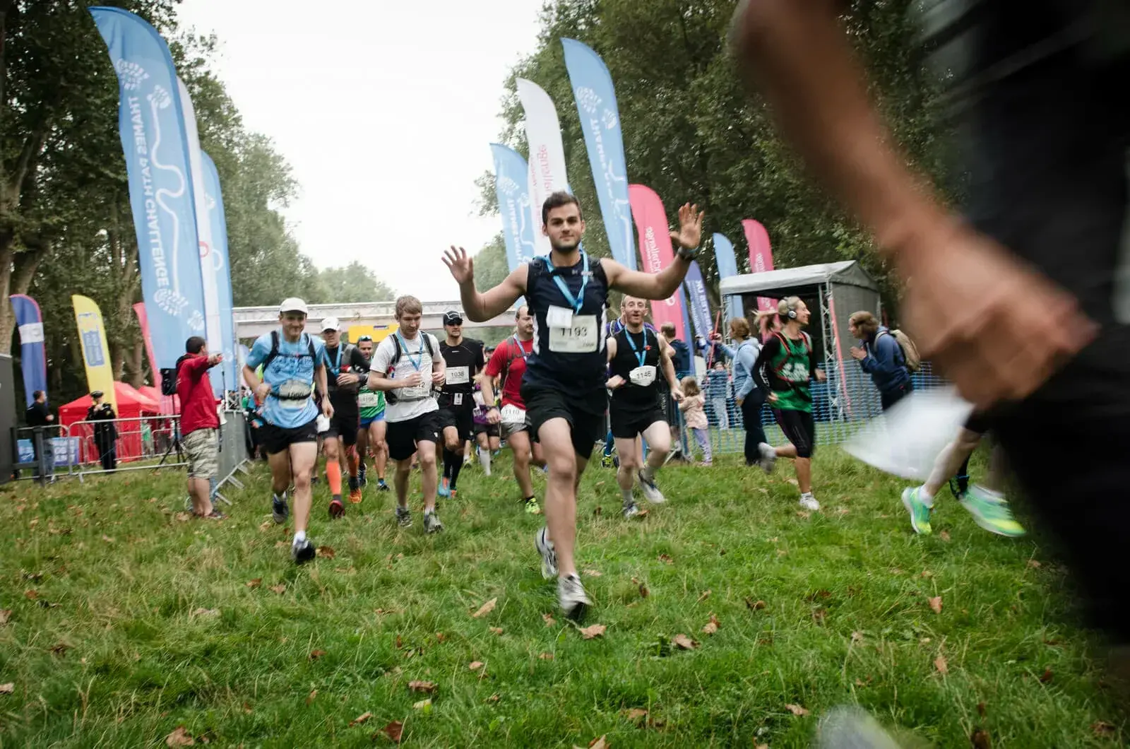 A group of runners participating in an outdoor race, with one man smiling and raising his arms. They are running on a grassy area, surrounded by trees and colorful flags. Spectators and event tents are visible in the background.