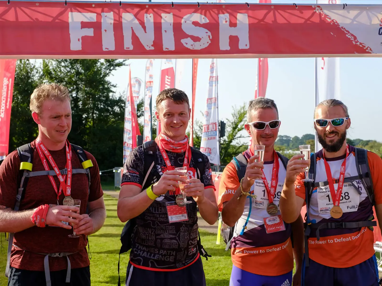 Four men wearing race bibs and medals stand under a finish line banner, smiling and holding drinks. They appear to have just completed a sports event, with trees and flags visible in the background.