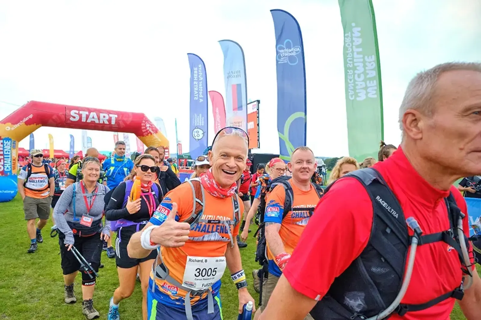 A group of people in athletic gear smiles as they participate in an outdoor event. Colorful flags and a "Start" arch are visible in the background. They are wearing numbered bibs with lanyards, and the atmosphere seems cheerful and lively.
