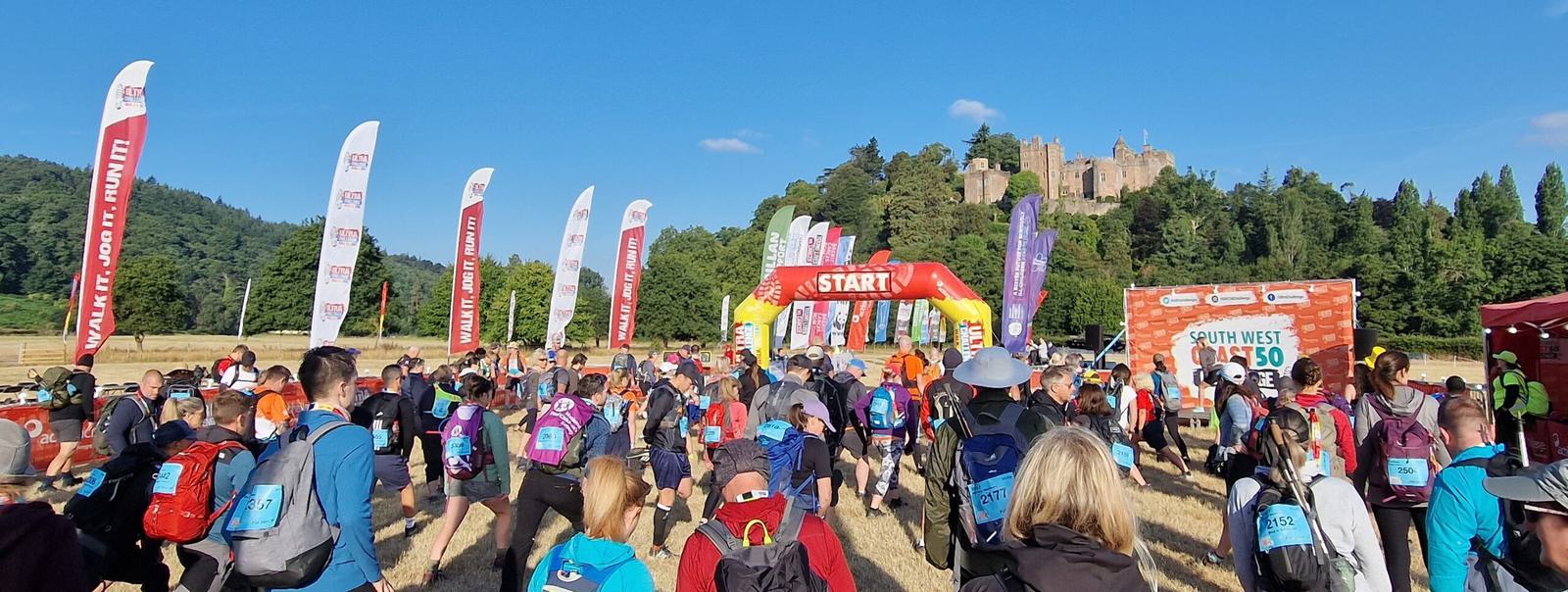 A large group of people, wearing backpacks and athletic gear, gather at the starting line of a race under a red arch labeled "START." Colorful flags line the path, and a historic castle is visible in the background against a clear blue sky.