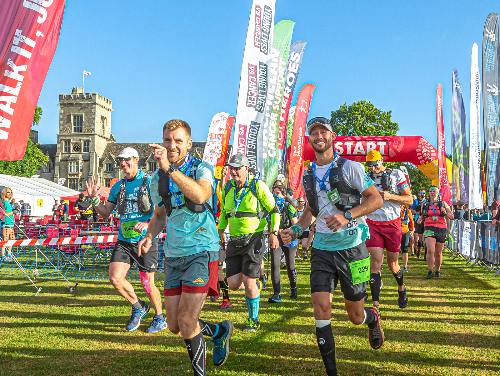 Runners in sporty attire participate in an outdoor event, smiling as they pass the start line. Colorful banners and a historic building are visible in the background, with bright blue skies above.