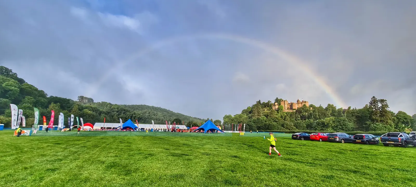 A large, grassy field with a rainbow arching across the cloudy sky. Several blue tents and parked cars are in the distance. A child in a yellow shirt runs on the grass, with a forested hillside and a castle visible in the background.