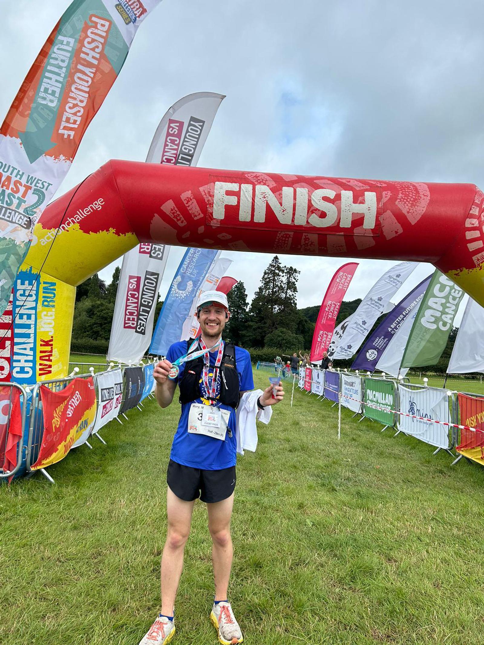 A runner stands smiling under a red "Finish" arch, holding a medal and a drink. He wears a race bib and is surrounded by colorful flags and banners on a grassy field, celebrating completing a race.