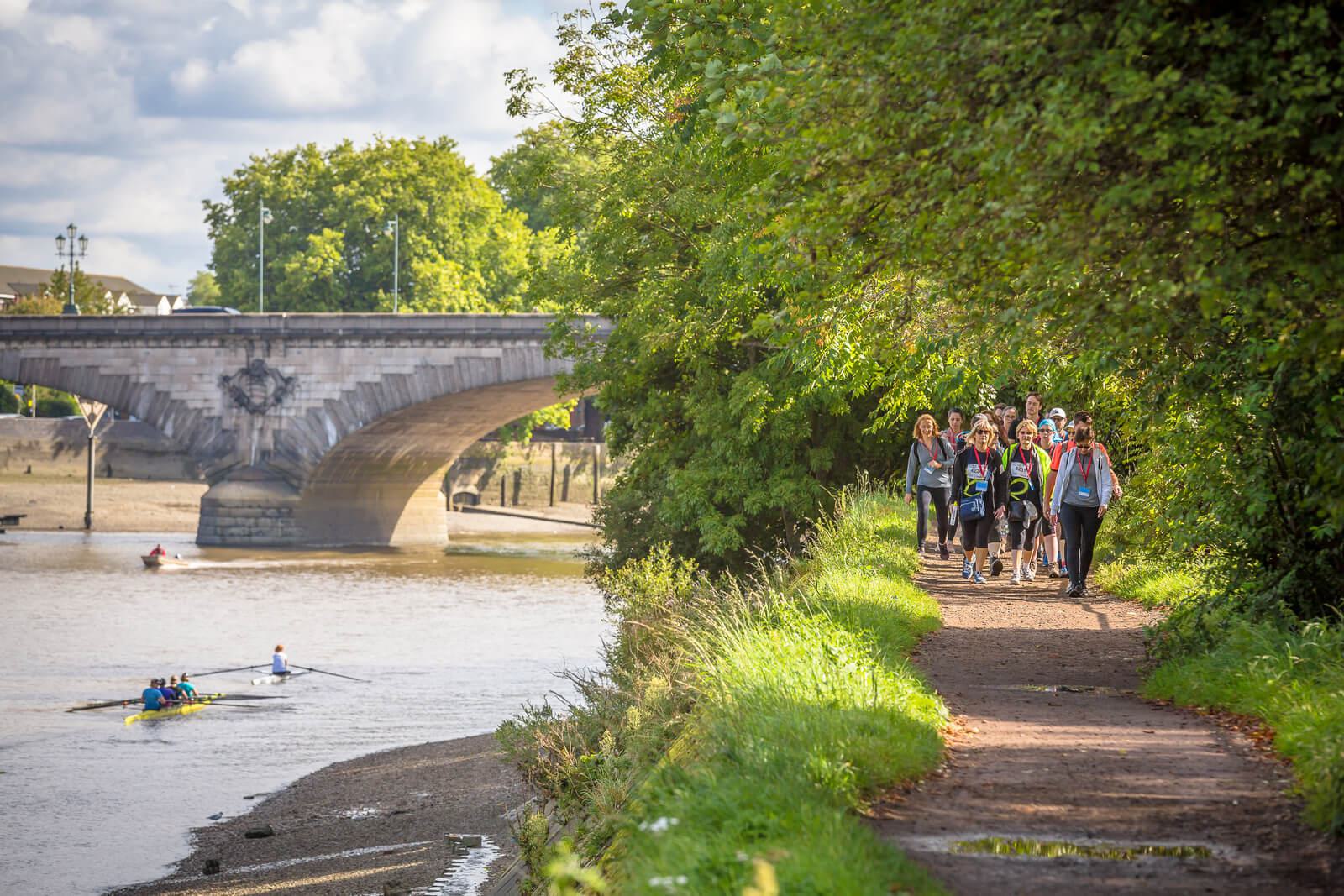 A group of people walks along a riverside path with lush greenery on one side. A rowboat with two people is in the river. A stone bridge arches over the river in the background under a partly cloudy sky.