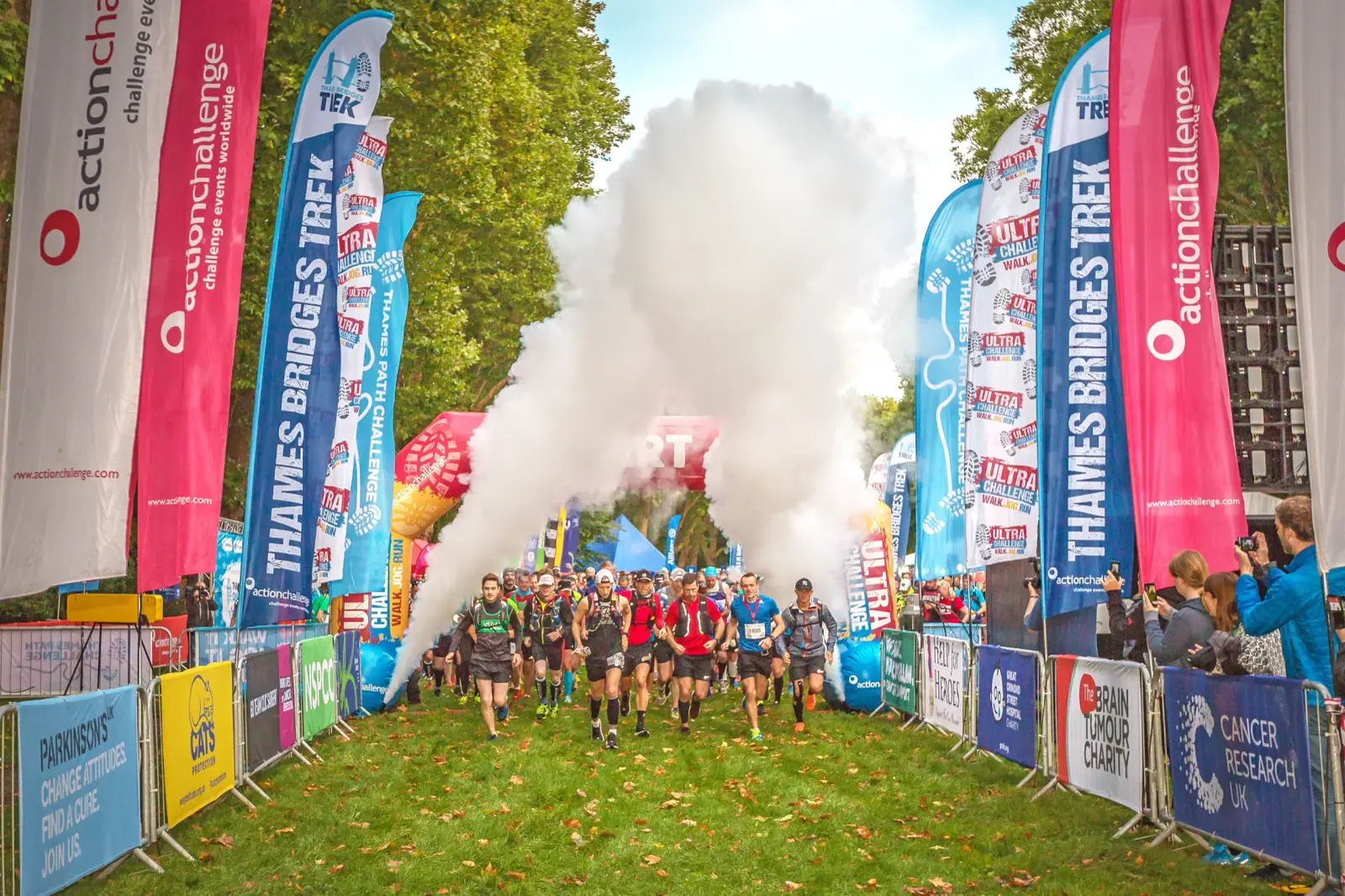 A group of runners wearing athletic gear start a race, passing under colorful banners with "Thames Bridges Trek" written on them. Smoke rises in the background, and spectators line the path, cheering on the participants.