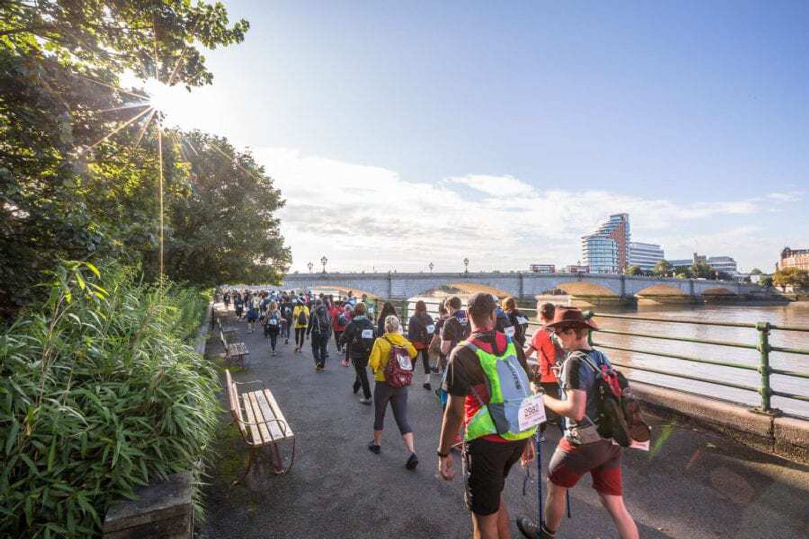 A large group of people walks along a riverside path under a clear blue sky. Sunlight filters through the trees on the left, casting shadows. A bridge and modern buildings are visible in the background.