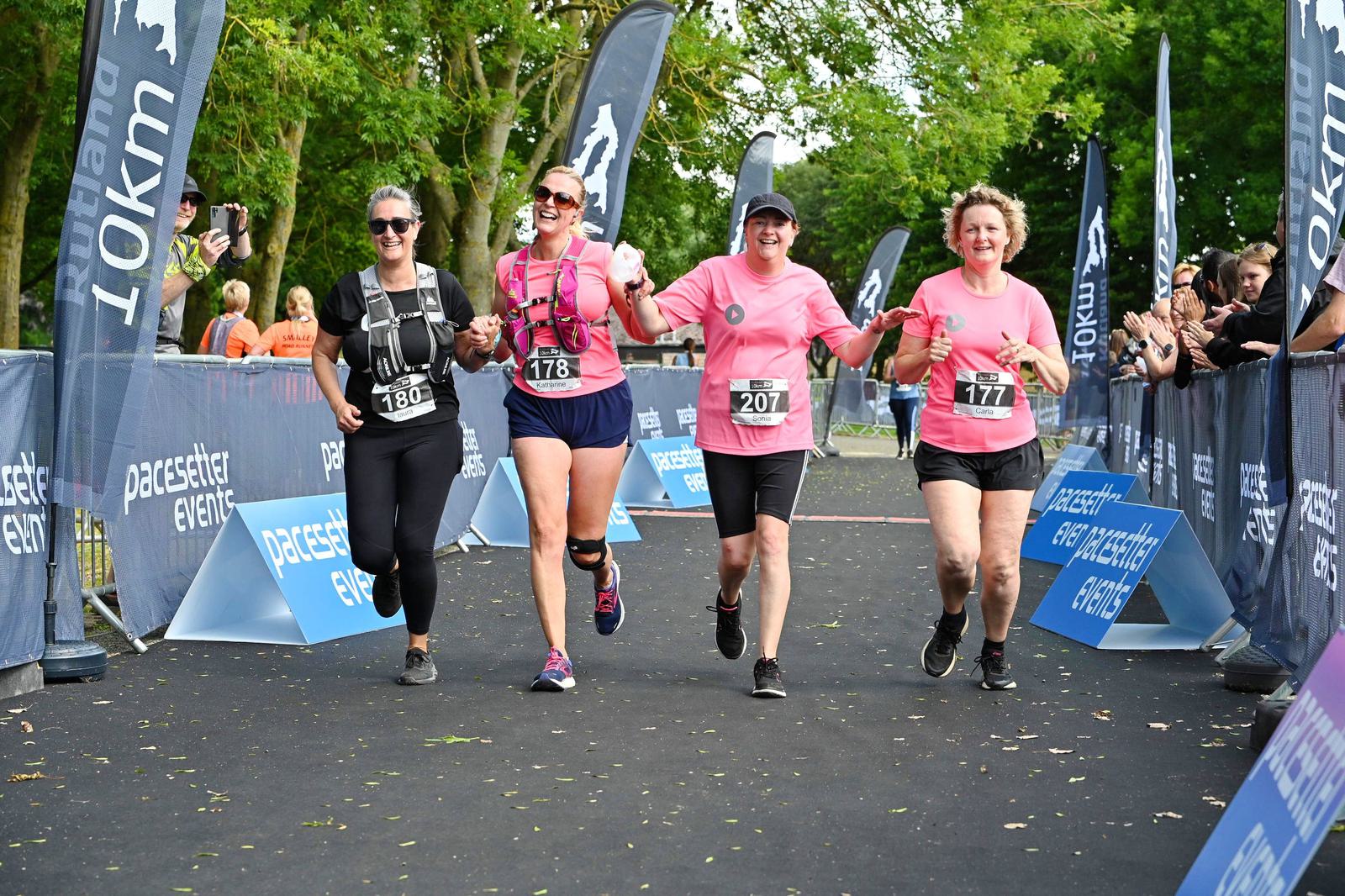 Four women holding hands and smiling while running towards the finish line of a race. They wear bib numbers and bright athletic clothing. A cheering crowd stands behind barriers, with banners displaying "pacesetter events" and "10km.