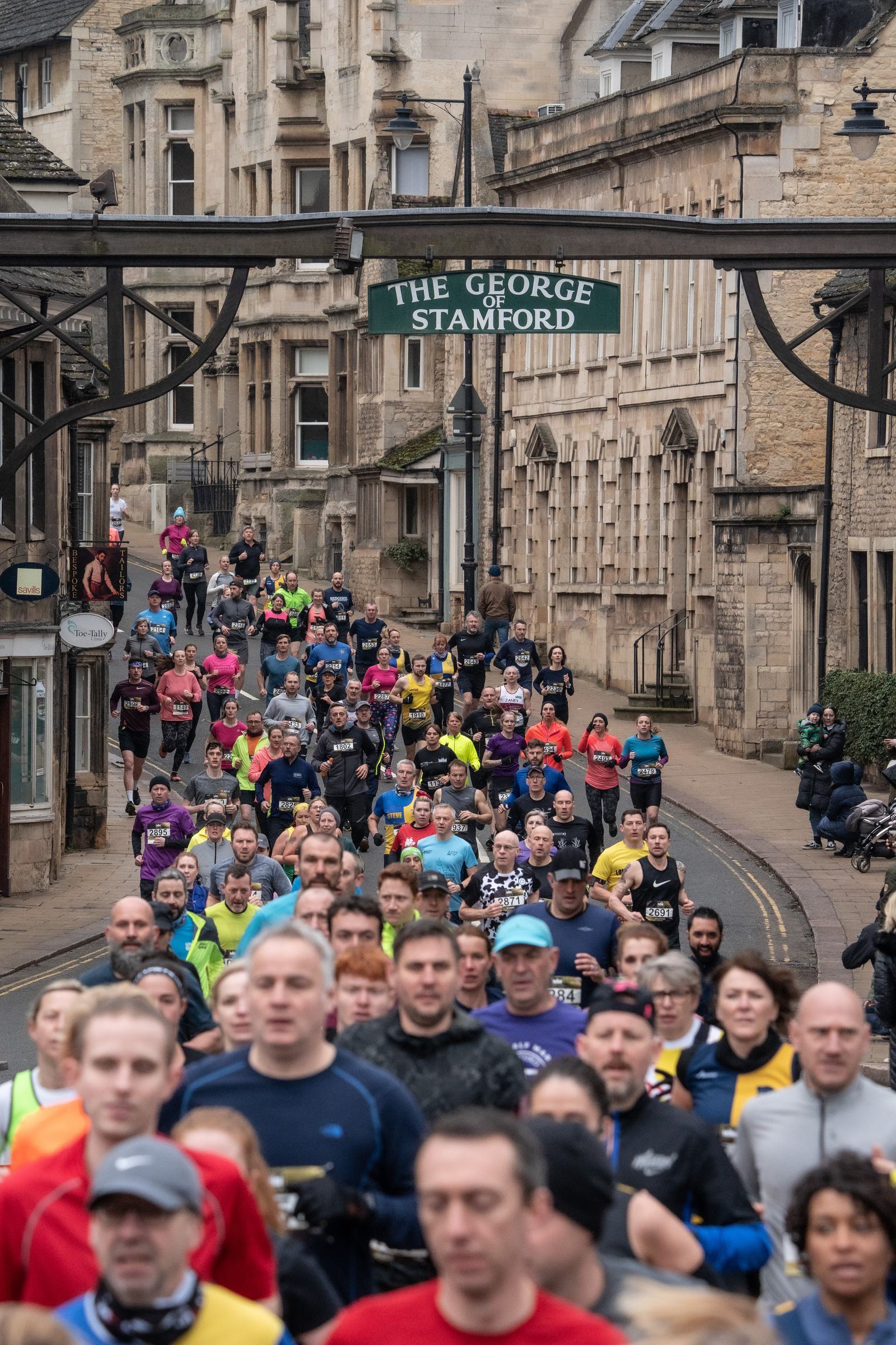 Crowd of runners participating in a street race in a historical town. Stone buildings line the road, and a sign reading "The George Stamford" hangs overhead. Participants wear various colorful athletic gear.
