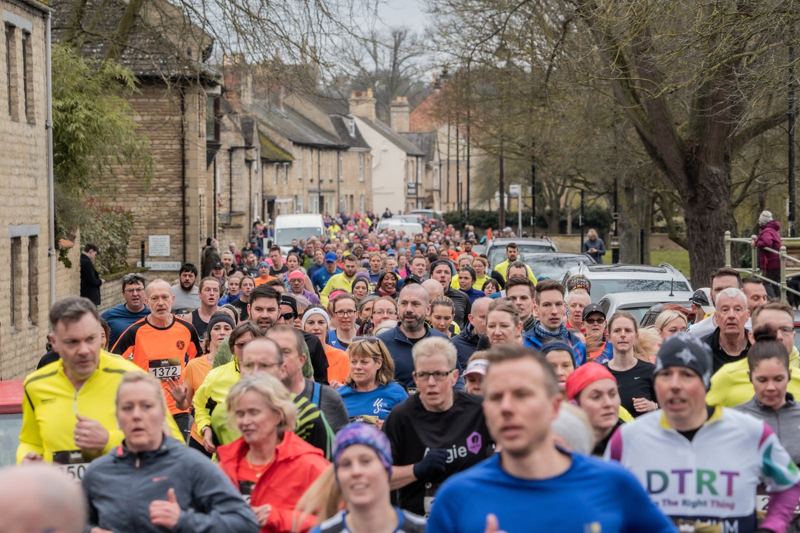 A large group of people running in a race on a street lined with trees and houses. Many participants are wearing colorful athletic clothing and some have numbers on their shirts. The atmosphere is lively and energetic.