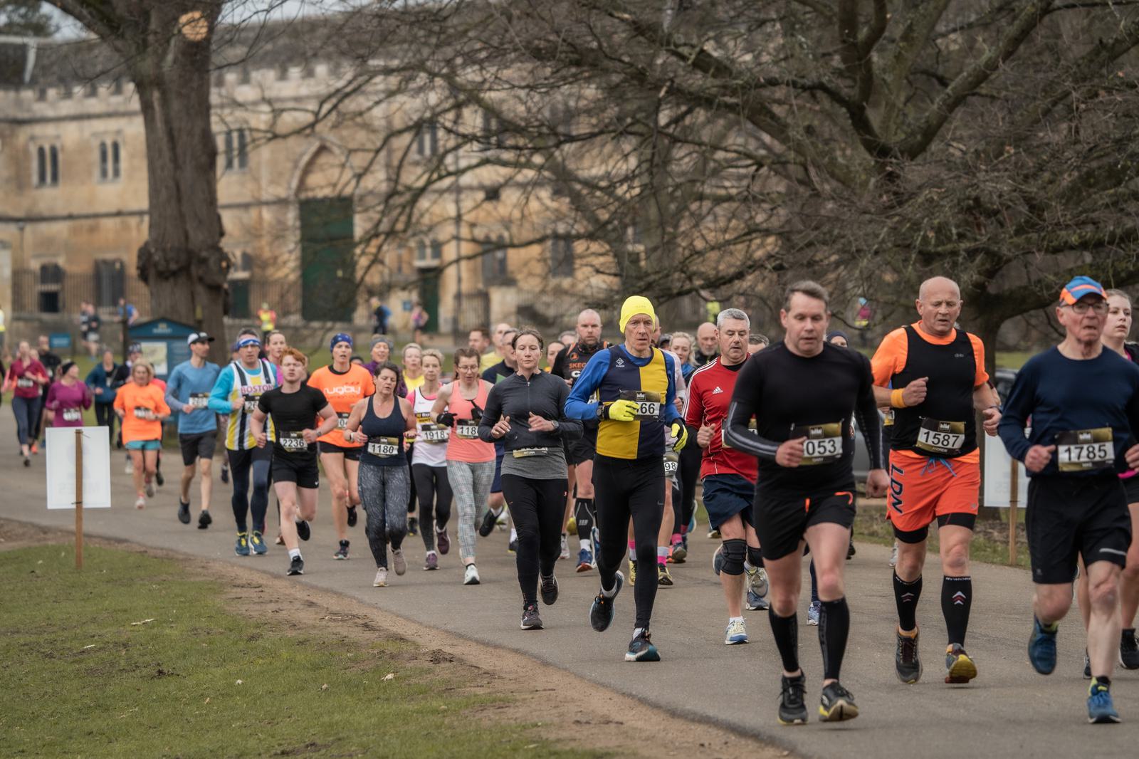 A group of runners in athletic gear participating in a race on a paved path. The background features trees and a historic stone building, creating an outdoor setting. The runners are diverse in attire and focused on the course.