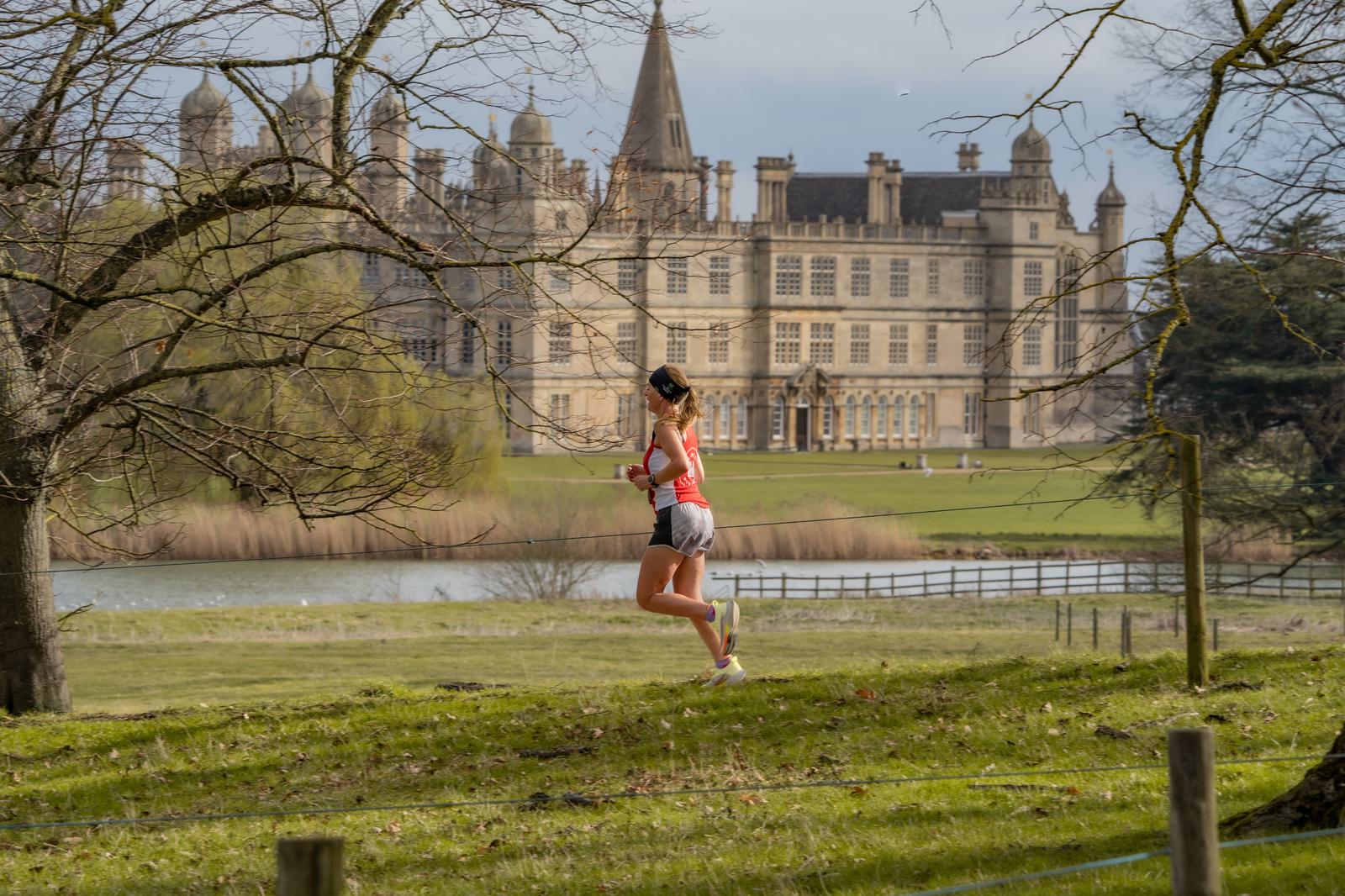 A woman in athletic gear jogs through a grassy park with trees. In the background, there's a large, historic building with towers and intricate architecture, set beside a pond. The sky is clear, creating a serene, sunny atmosphere.