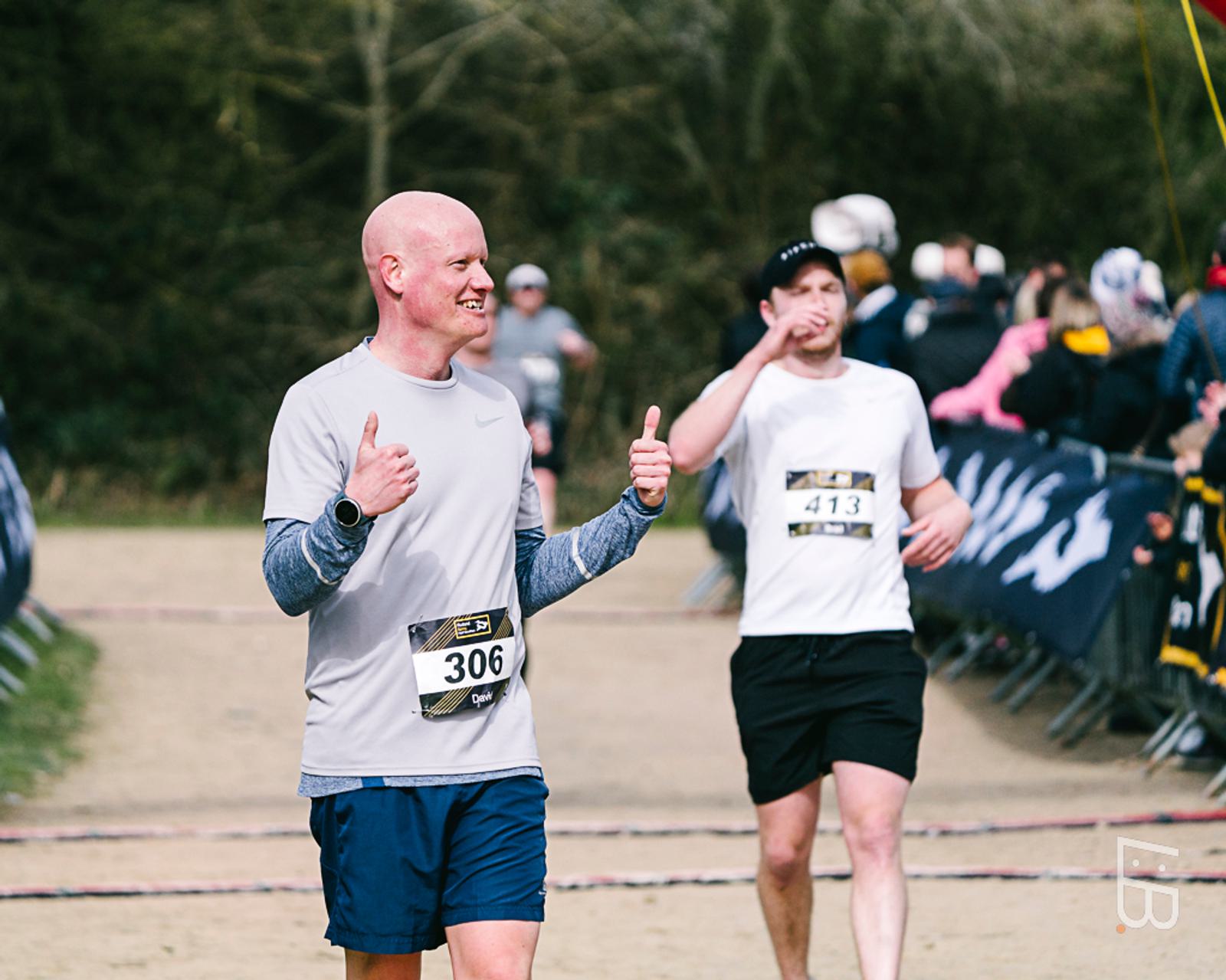 A man in a gray shirt and numbered bib gives two thumbs up as he finishes a race. Behind him, another runner in a black and white outfit approaches the finish line. Spectators watch from the sidelines.