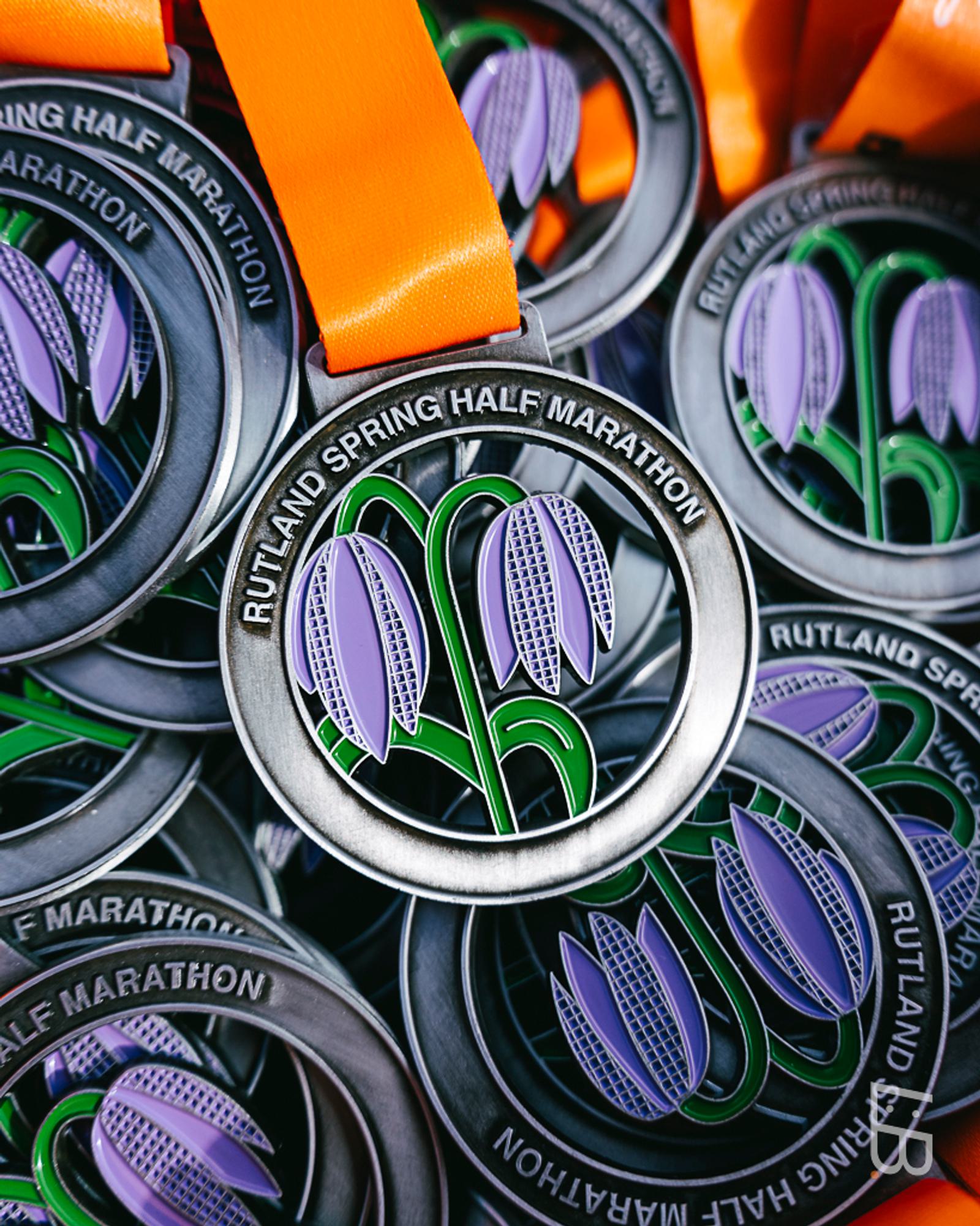 Close-up of Rutland Spring Half Marathon medals with orange ribbons. The medals feature an intricate design of purple flowers and green stems, with the event name engraved around the edge.