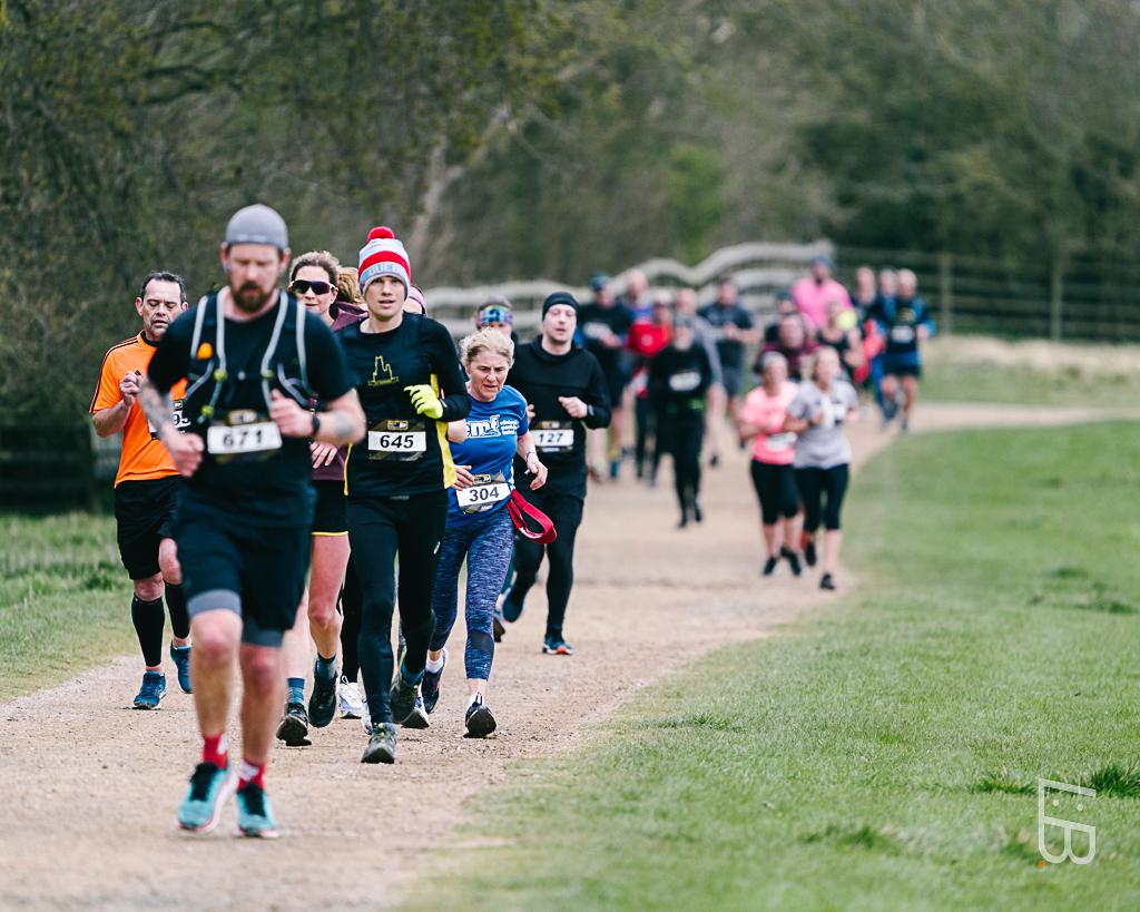 A group of runners participating in a race on a gravel path through a park. The lead runner, wearing a gray headband and number 671, is followed by others, some in colorful attire. The setting is lush and green, with trees lining the path.