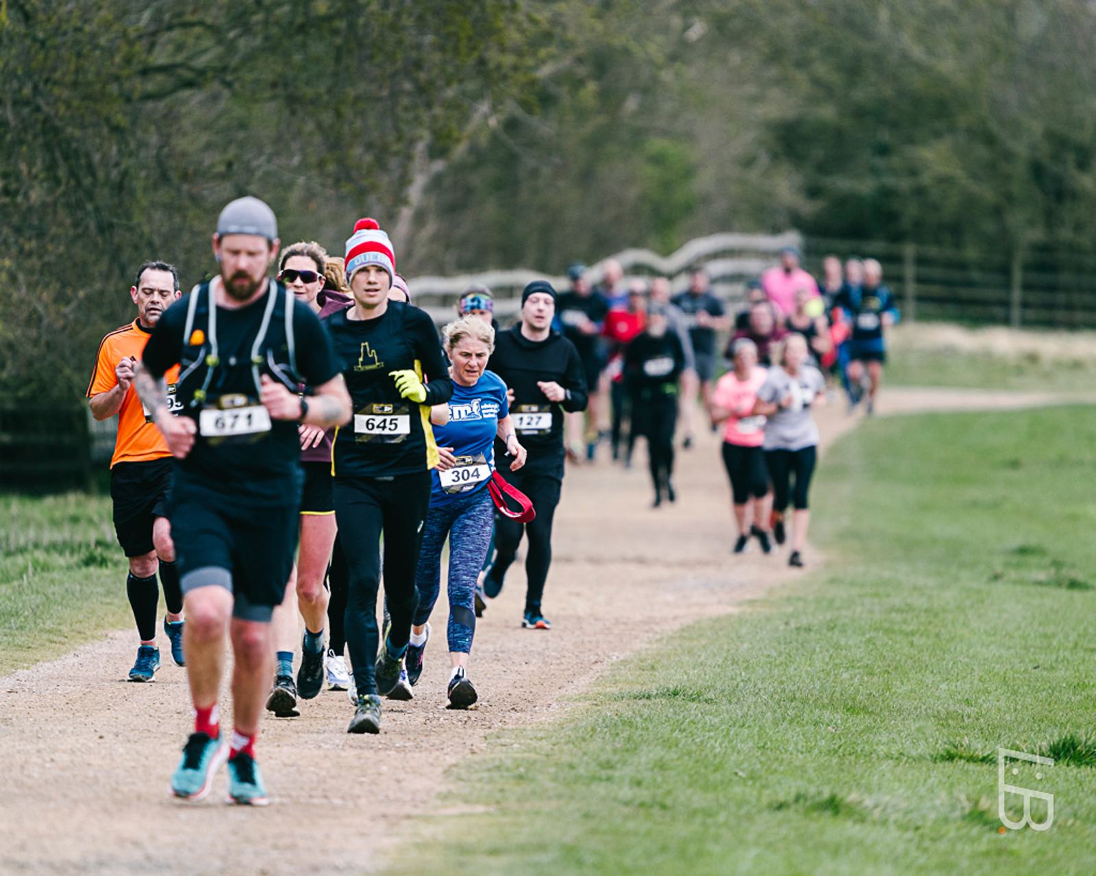 A group of runners participating in a race on a gravel path through a park. The lead runner, wearing a gray headband and number 671, is followed by others, some in colorful attire. The setting is lush and green, with trees lining the path.
