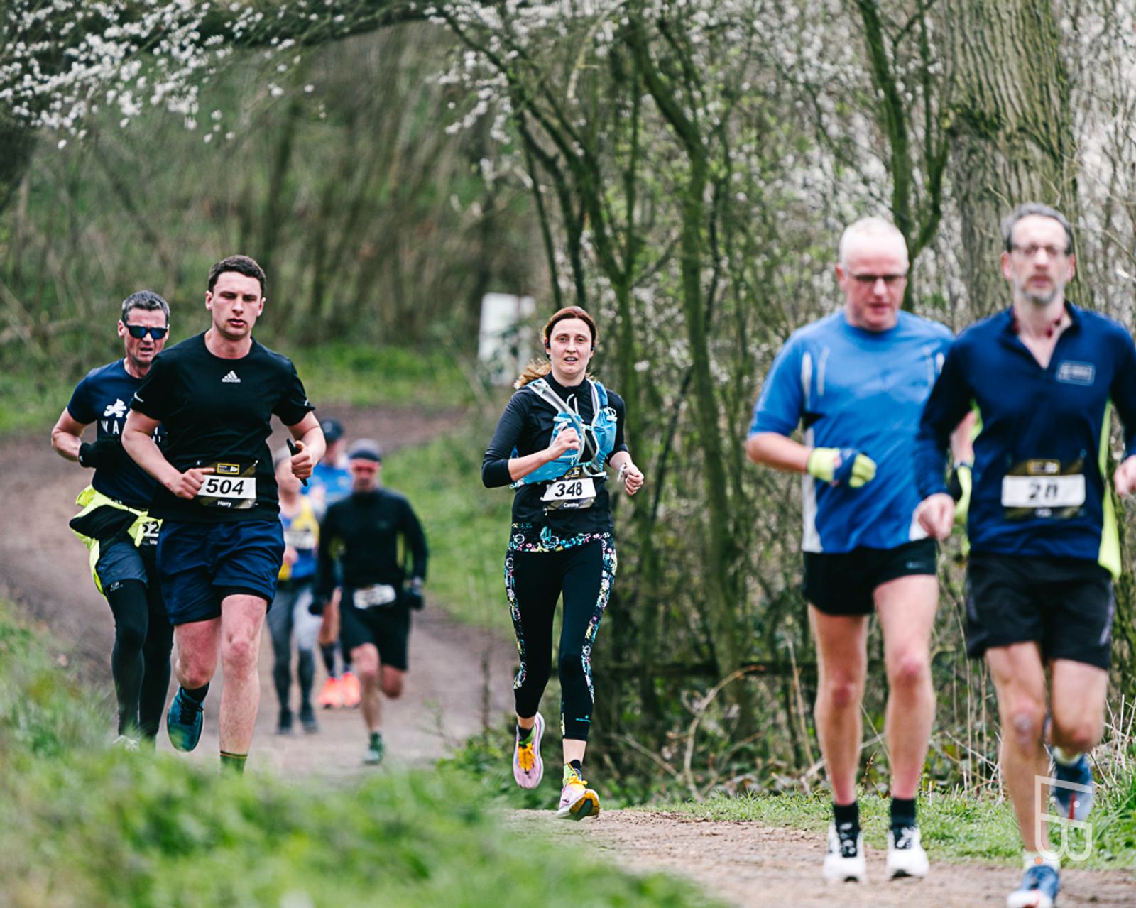 A group of runners in a race on a dirt path surrounded by trees. They wear different athletic outfits and race numbers. The background features a lush, green landscape, indicating a likely spring setting.