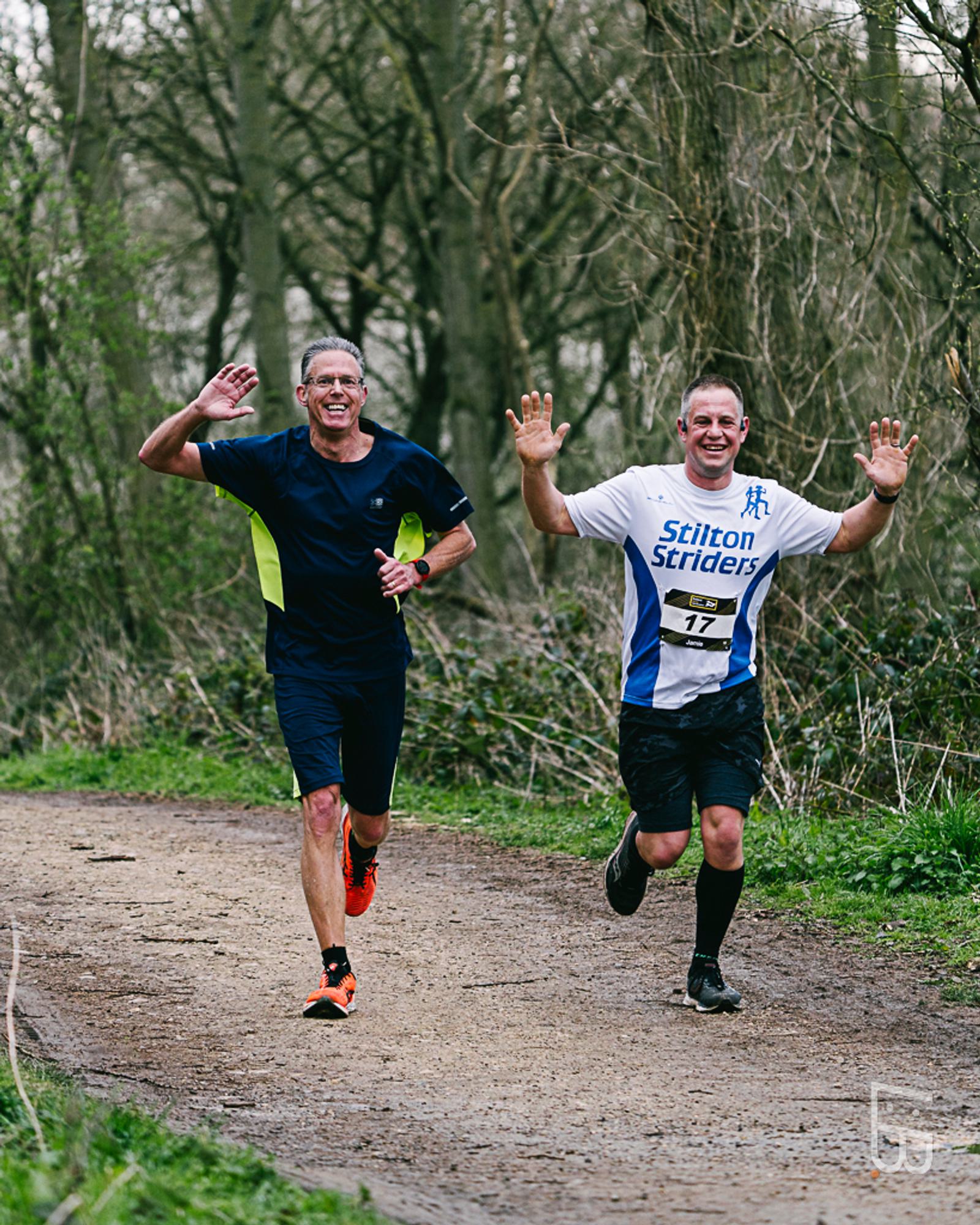 Two runners smiling and waving at the camera while jogging on a forest trail. One wears a dark athletic outfit; the other wears a "Stilton Striders" shirt with the number 17. Both seem energized and happy. Trees and greenery surround the path.