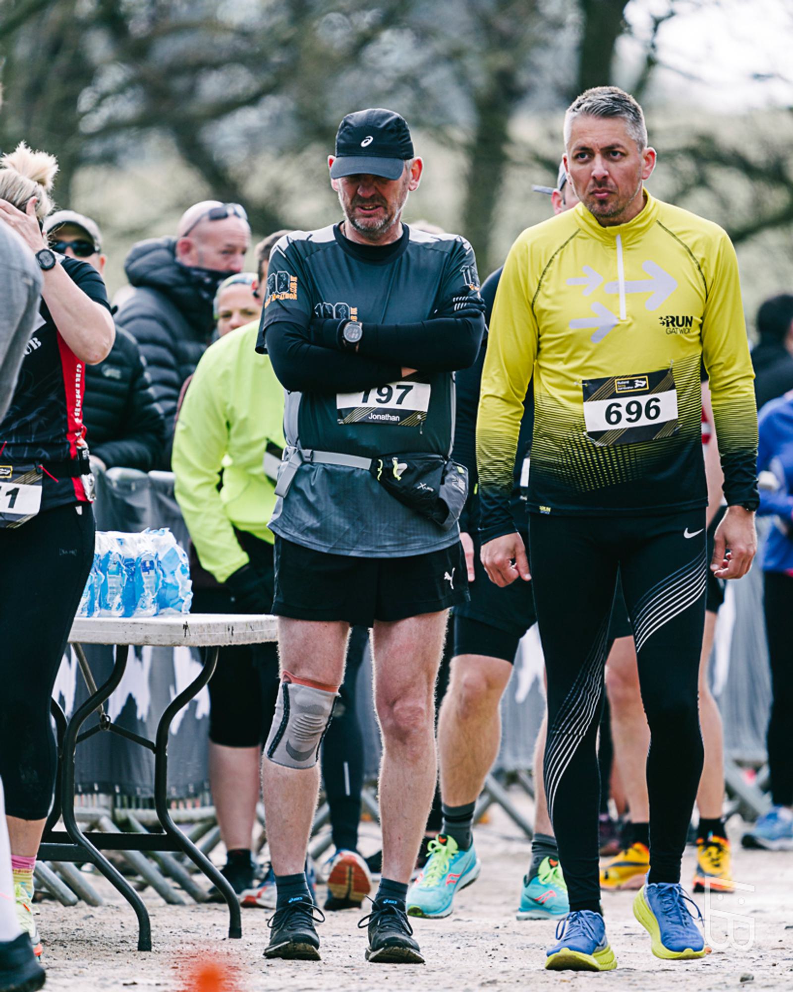 Two men in athletic gear stand at a race event. The man on the left wears a black cap and knee brace. The man on the right wears a yellow shirt with number 696. Other participants and a table with water bottles are visible in the background.