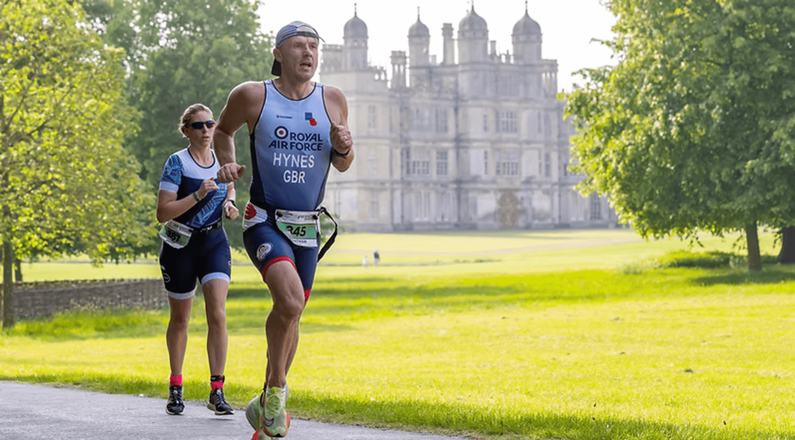 Two athletes in race gear, one in a blue tri-suit with "Royal Air Force" text, run along a path in front of a historic building. The background has lush green trees and grass, with the sun shining brightly.